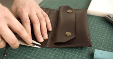 Craftsman pulling thread to tighten stitches while hand sewing leather wallet. Detail of handmade leatherworking technique.