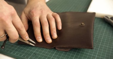 Craftsmans hands shaping brown leather wallet using professional leather tools. Close-up emphasizes artisanal skills, precision, and handmade production.