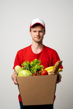 Smiling delivery man wearing red uniform and cap holding cardboard box with vegetables and fruits. Concept of healthy food delivery service, courier, organic groceries and urban logistics.
