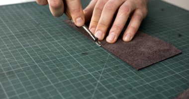 Close-up of craftsman hands using edge tool to process leather strip on cutting mat during handmade leather goods production.