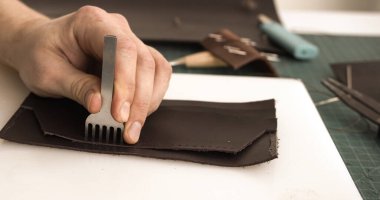 Artisan hand punching stitching holes in leather piece with pricking iron tool on cutting mat for handmade leather goods.