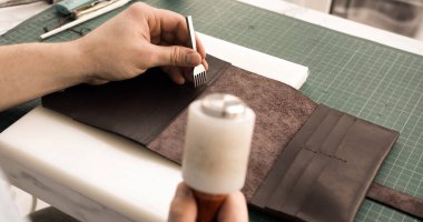 Close-up of leatherworking process with tools and dark leather. Craftsman uses awl and mallet for precise stitching preparation. Concept of craftsmanship, workshop, artisan handmade work.