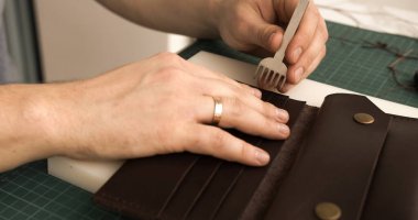 Close-up of punching stitching holes in a handmade leather wallet. Artisan uses pricking iron and tools for precise stitching preparation. Concept of craftsmanship and handmade work.
