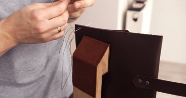 Artisan sewing leather manually using thread and needle fixed on wooden stitching pony. Detail of handmade leather crafting.