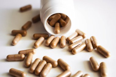 Beyaz arka planda beyaz plastik şişeden dökülen kahverengi bitkisel kapsüller doğal katkı maddelerini, vitaminleri, sağlık hizmetlerini ve besinleri sembolize ediyor..