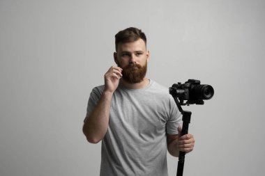 Bearded man in grey t-shirt operating professional camera on gimbal stabilizer against plain background, symbolizing filmmaking, video production.