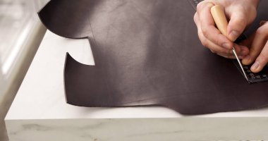 Detail of craftsman hands marking genuine leather with ruler and awl for precise handmade leather goods production in workshop.
