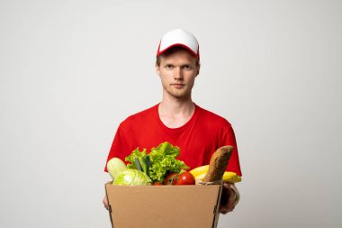 Delivery man in red t-shirt and cap holding grocery box with bread, bananas, tomatoes, lettuce, and cabbage. Concept of online shopping, courier service, and food delivery.