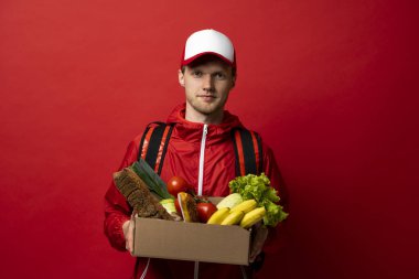 Male courier in red cap and jacket carrying grocery box with bananas, bread, lettuce, and tomatoes. Concept of online shopping, food delivery, courier service, and logistics.