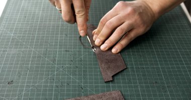 Hands cutting leather strip with knife on green cutting mat. Concept of handmade leatherwork, craftsman skills, artisan production, and material design.