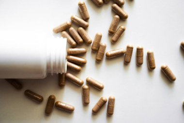 Kahverengi bitkisel kapsüller beyaz bir yüzeyde beyaz bir plastik şişeden döküldü. Sağlık hizmetleri, doğal takviyeler, vitaminler, alternatif tıp ve sağlığın sembolü.