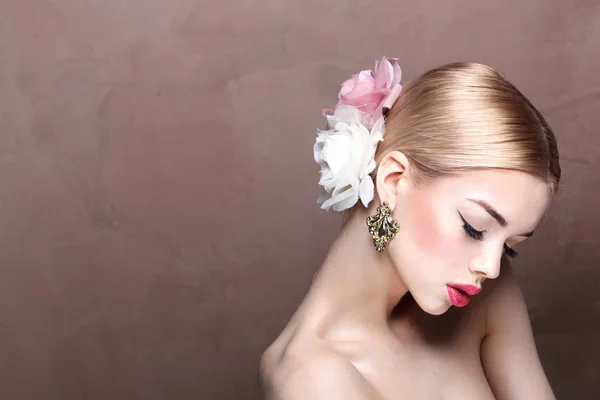 young blonde woman with pink lips and roses in her hair wearing vintage earrings posing on brown background with copy space