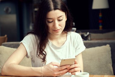 Ahşap masa kahve dükkanında otururken, kahve içme ve smartphone kullanarak genç kadın. Beyaz fincan espresso masada duruyor.