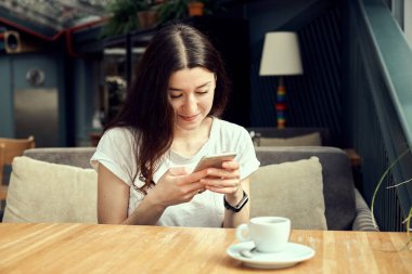 Ahşap masa kahve dükkanında otururken, kahve içme ve smartphone kullanarak genç kadın. Beyaz fincan espresso masada duruyor.
