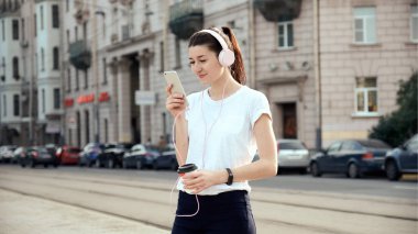 Akıllı telefon, müzik kulaklık açık havada kullanan genç ve güzel kadın yemek kahve fincanı, içme. Kentsel yaşam tarzı kavramı. Sağlıklı, pozitif, neşeli sabah çalışan.