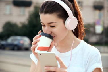 Akıllı telefon, müzik kulaklık açık havada kullanan genç ve güzel kadın yemek kahve fincanı, içme. Kentsel yaşam tarzı kavramı. Sağlıklı, pozitif, neşeli sabah çalışan.