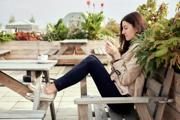 若い女性のコーヒー ショップで木のテーブルに座って コーヒーを飲みながら スマート フォンを使用しています エスプレッソの白いカップがテーブルの上に立つ — ストック写真