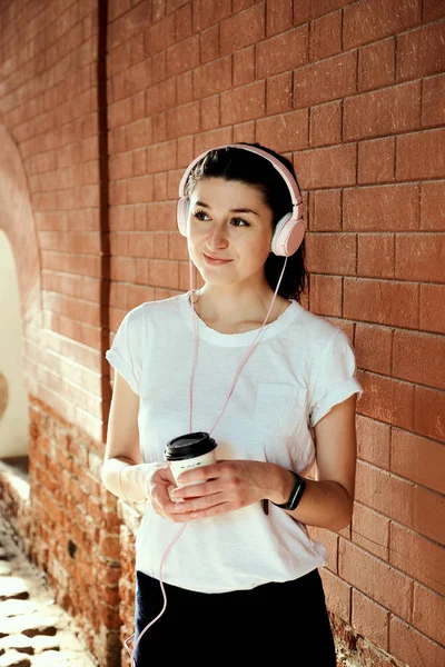 Young pretty caucasian woman with coffee cup in her hands stand alone near brick wall. Early morning running, wake up, listen to music, fashion girl, sweet drink, smoothie health, aromatic coffee