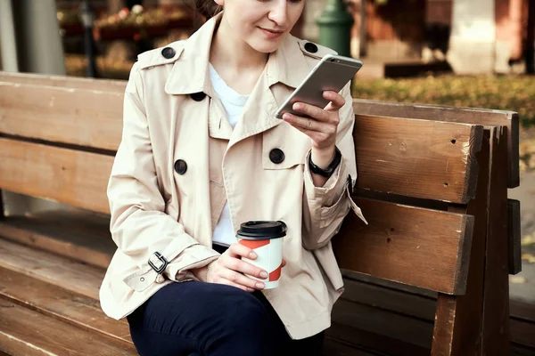 美しい若い女性は木製のベンチに座っている コーヒーのテイクアウトのカップを保持するスマート フォンを使用して — ストック写真