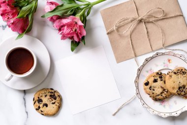 Fincan kahve, pembe çiçekler, kartpostal, kraft zarf, sicim, pamuk şube, yulaf kurabiye, metin kopya alanı ile davetiye kartı ile mermer masası. Düz yatıyordu, en iyi görünüm. Kadının gün ya da anneler günü.