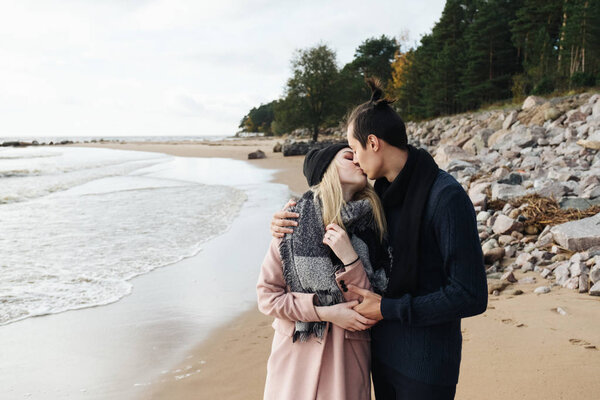 Couple in love walking on the beach, hugging and kissing. Autumn weather, coastline and forest on the background. Young man wearing a wool sweater, girl in a pink coat.