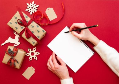 Kadınların elleri kalem ve defter dilek listesi ile yılbaşı süsleri ve hediyeleri kırmızı zemin üzerine tutarak. Noel ve mutlu yeni yıl kompozisyon. Yat style, üstten görünüm, tepegöz, alanı Kopyala