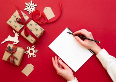 Kadınların elleri kalem ve defter dilek listesi ile yılbaşı süsleri ve hediyeleri kırmızı zemin üzerine tutarak. Noel ve mutlu yeni yıl kompozisyon. Yat style, üstten görünüm, tepegöz, alanı Kopyala