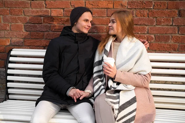 Happy caucasian couple in love in park sitting on the bench near brick wall with takeaway drink