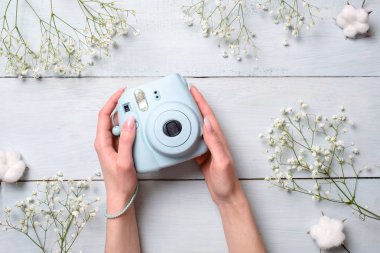 Çiçekli mavi ahşap arka plan üzerinde modern polaroid kamera tutan eller. Üstten Görünüm, ihale en az düz stil kompozisyon yatıyordu. Kadın Danışma, moda blogger, güzellik ve teknoloji kavramı