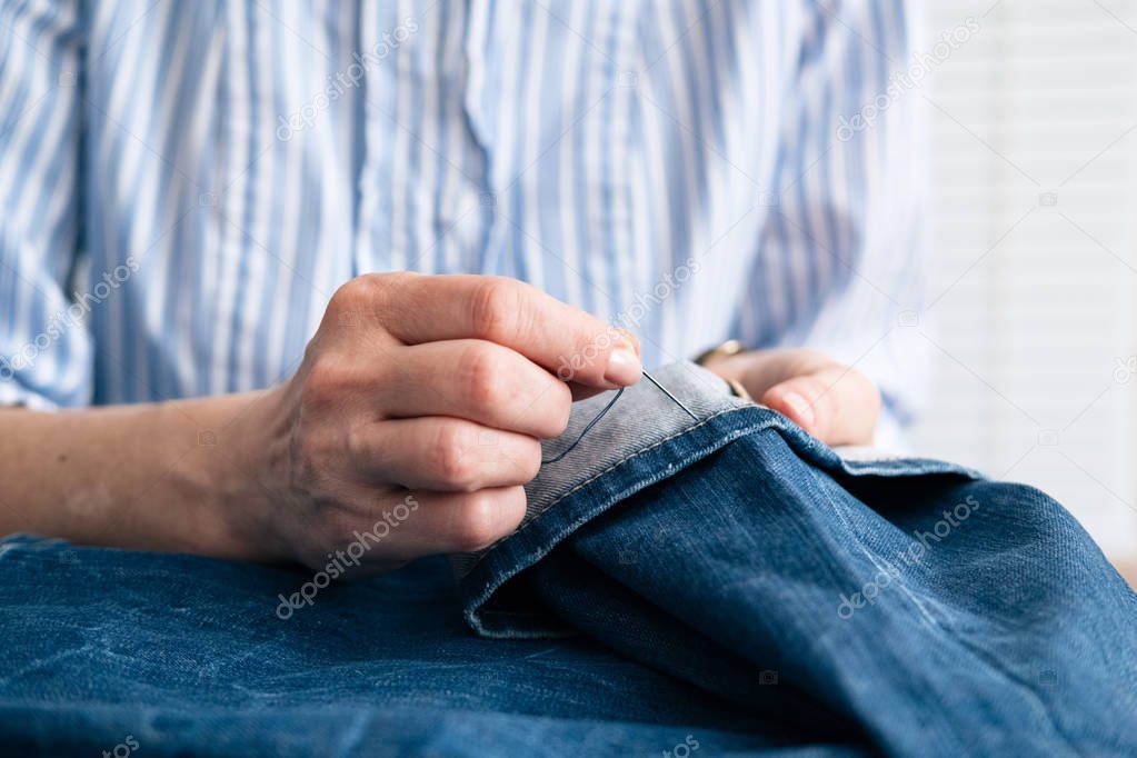 Tailor sew the hem of jeans in workshop, close up view on hands with sewing needle