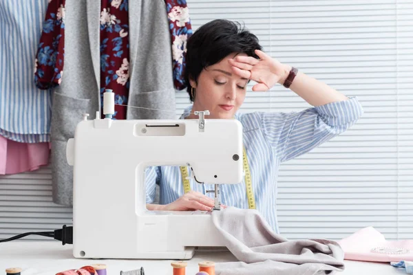 Frustrated tired seamstress touching her head, feeling absolutely exhausted because of overwork, working at tailor shop with electrical industrial sewing machine