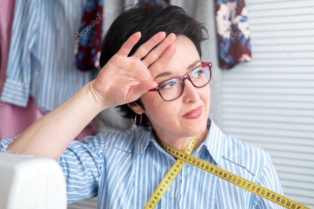 Frustrated tired seamstress touching her head, feeling absolutely exhausted because of overwork, working at tailor shop with electrical industrial sewing machine