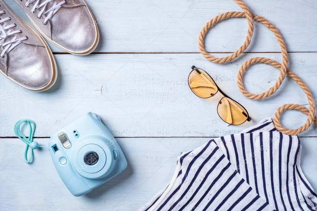 Frame from girls accessories on pastel blue wooden desk: instant photo camera, sunglasses, t-shirt.  Summer background, travel or vacation concept,  flat layout design, view from above.
