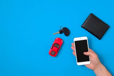 Araç Kiralama uygulaması konsepti. Oyuncak araba, Araba anahtarı, otomatik sürücü lisansı, mavi arka planda insan el tutan akıllı telefon. Düz bir kompozisyon. Üst görünüm.