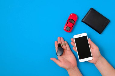 Araç Kiralama uygulaması konsepti. Oyuncak araba, otomatik sürücü lisansı, mavi arka plan üzerinde akıllı telefon ve araba araba anahtarı tutan insan elleri. Düz bir kompozisyon. Üst görünüm.