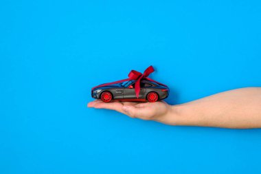 Oyuncak araba modeli kırmızı bir kurdele ile bağlı ve mavi arka planda insan eline yay. Otomatik Bayilik ve Kiralama, yeni oto satın, hediye veya mevcut olarak araba, araba çizmek, modern araba kavramı kazanmak için şans.