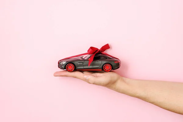 Human hand holding toy car model tied with a red ribbon and bow on pink background. Auto dealership and rental, car as gift or present, draw car, chance to win modern car concept — Stock Photo, Image