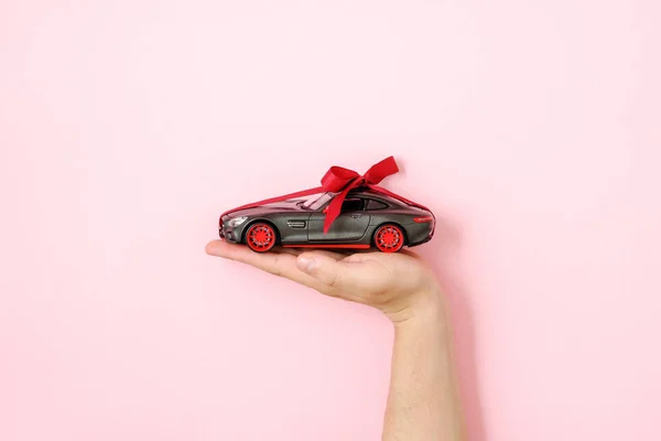 Toy car model with ribbon bow on human hand on pink background. Auto dealership and rental concept — Stock Photo, Image