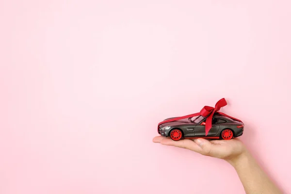 Toy car model tied with a red ribbon and bow on human hand on pink background. Auto dealership and rental, car as gift or present, draw car, chance to win modern car concept.