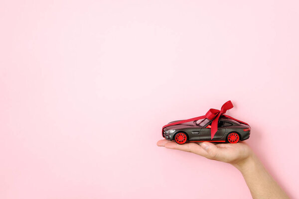 Игрушечная модель автомобиля привязана красной лентой и бантиком на человеческой руке на розовом фоне. Автосалон и прокат автомобилей, автомобиль в подарок или подарок, привлечь автомобиль, шанс выиграть современную концепцию автомобиля
.