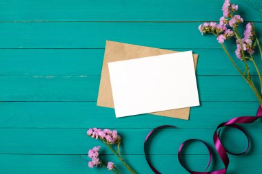 Yeşil ahşap masa üzerinde mor papatya çiçekleri ile boş tebrik kartı ve zarf. Düz döşeme kompozisyonu, üst görünüm, genel merkez.