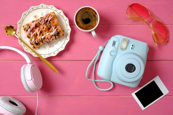 Composition with instant camera, picture frame, headphones, cup of coffee tasty wafer cake on pink wooden background. Beauty blogger concept. Stylish hipster women desk. Flat lay. Top view.