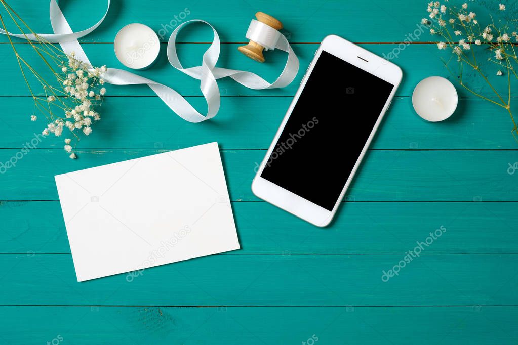 Beauty blogger woman's desk concept. Flat lay style composition with modern smartphone, daisy flowers, ribbon and blank paper card on green wooden background. Banner mockup for beauty salon.
