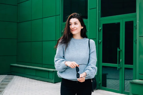 Mulher bonita em um suéter de lã azul posando na rua da cidade no fundo urbano edifício verde. Senhora de negócios confiante, menina hipster elegante na roupa da moda com óculos de sol em sua mão . — Fotografia de Stock