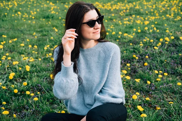Close up portrait of beautiful millennial young woman in sunglasses sitting on meadow with yellow chamomile flowers and grass in park. Natural beauty and relaxation concept. — Stock Photo, Image