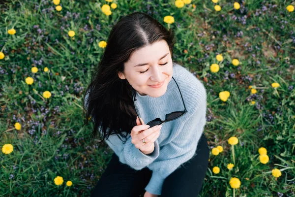 Primer plano retrato de hermosa joven milenaria en gafas de sol sentado en el prado con flores de manzanilla amarilla y hierba en el parque. Concepto de belleza natural y relajación . — Foto de Stock