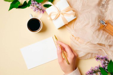 Üst görünüm, düz yatıyordu kadın masası. Leylak çiçekler, ipek eşarp, parfüm şişesi, hediye kutusu ve bej kadınsı arka plan üzerinde boş tebrik kartı. Zarif ve çekici kadın şeyler. Düğün davetiyesi konsepti.