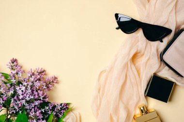 Bej arka plan üzerinde ipek eşarp, parfüm şişesi, güneş gözlüğü, hipster aksesuarları ve leylak çiçekleri ile kadınsı masa çalışma alanı. Üst görünüm, ihale minimal düz döşeme stil kompozisyon kopya alanı ile.