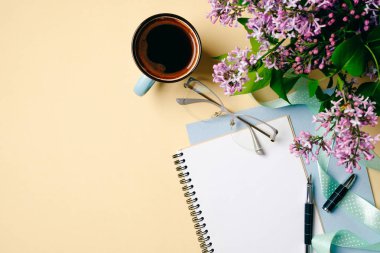 Düz yatıyordu ev ofis masası. Beyaz kağıt defter, kahve fincanı ve bej arka plan üzerinde leylak çiçekler ile Kadın çalışma alanı. Üst görünüm kadınsı arka plan. Moda blogger masaüstü konsepti.