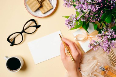 Düz yatıyordu ev ofis masası. Boş kağıt kartı ile Kadın çalışma alanı, el tutma kalemi, hediye kutusu, kahve fincanı, waffle, bej arka plan üzerinde leylak çiçekler. Üst görünüm kadınsı arka plan. Moda blog kahraman.